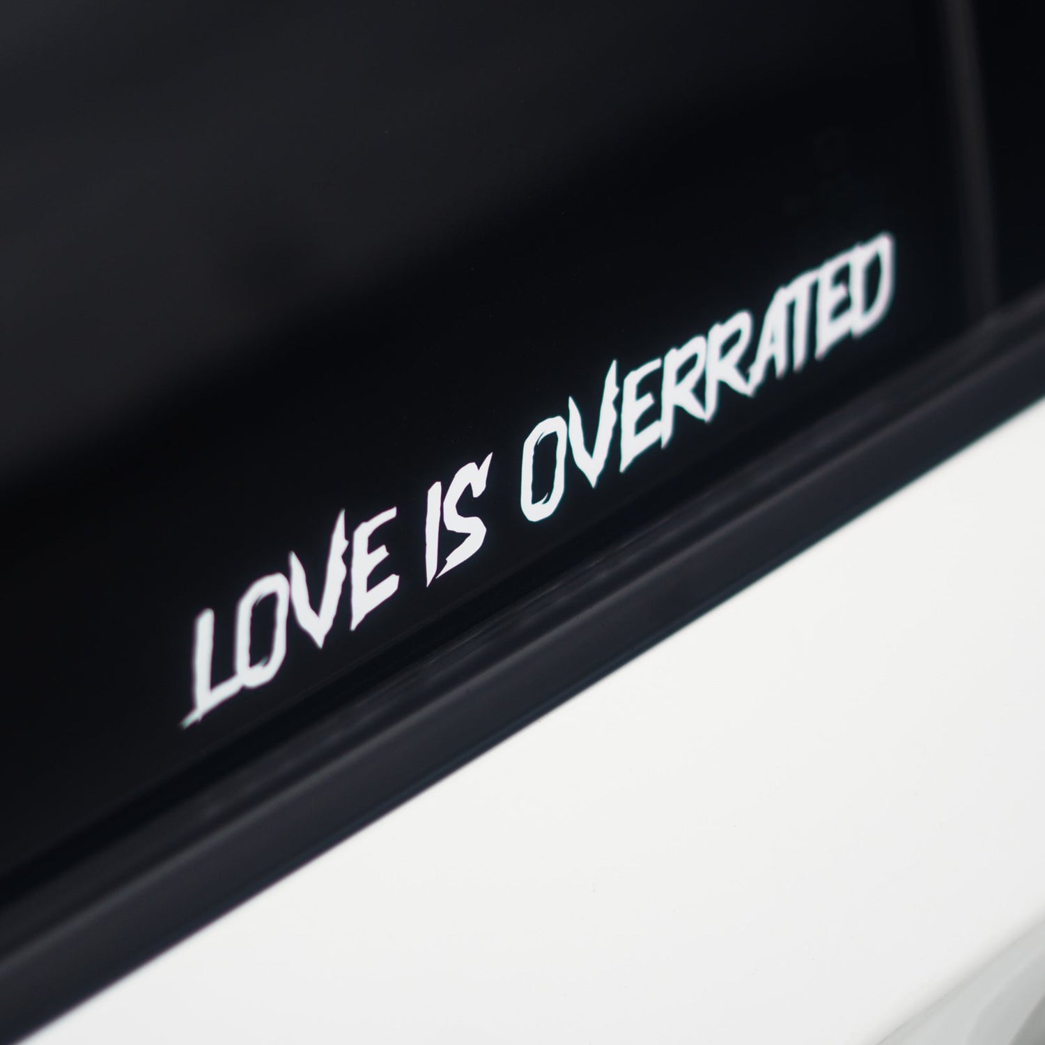 A white vinyl decal sticker with the text 'LOVE IS OVERRATED' in a stylized font, applied to the black window trim of a white car.